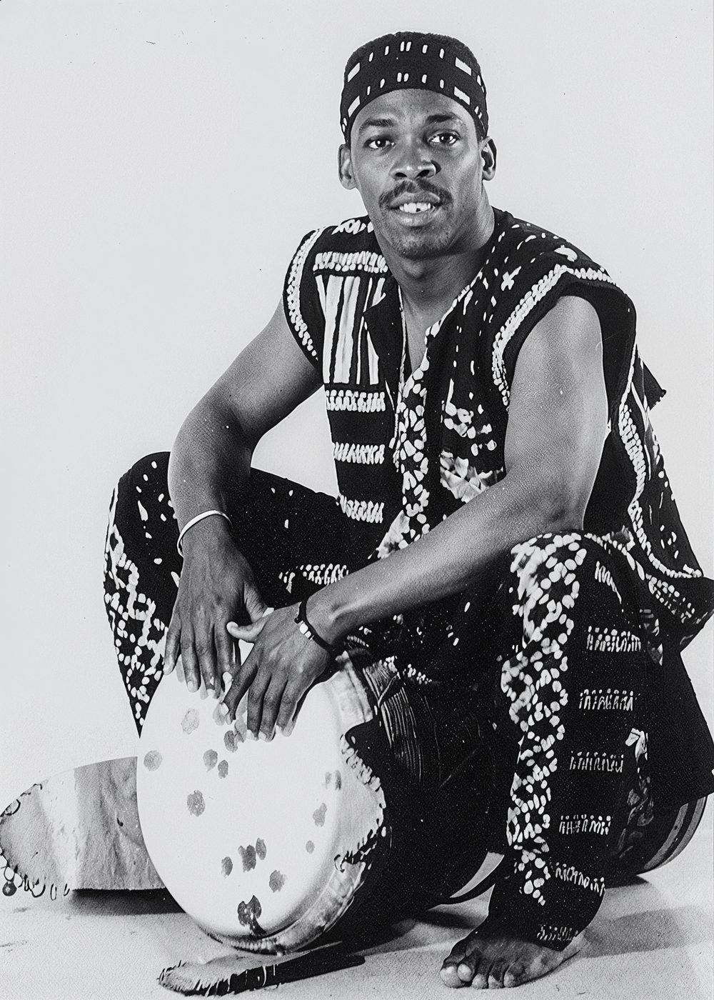 Percussionniste accroupi en tenue à motifs tenant un djembé, photographie en noir et blanc